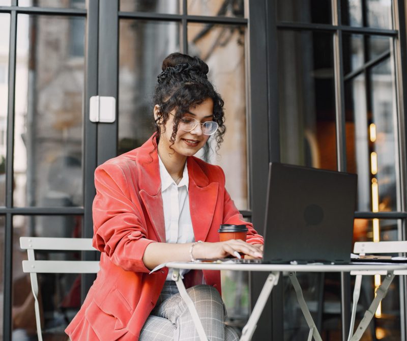 Indian woman working on a laptop in a street cafe. Wearing stylish smart clothes - jacket, glasses
