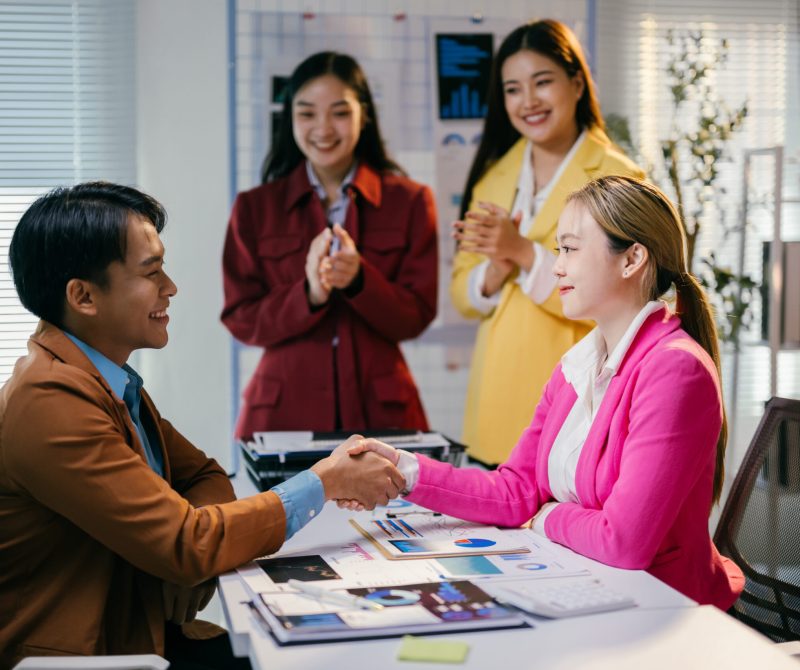 Young businessman and businesswoman shaking hands after closing a deal in a modern office, their teammates are applauding them in the background