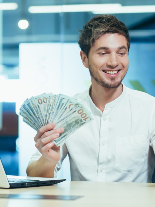 Young successful freelancer in the office holding in his hand winning banknote dollars, smiling and looking at the phone screen