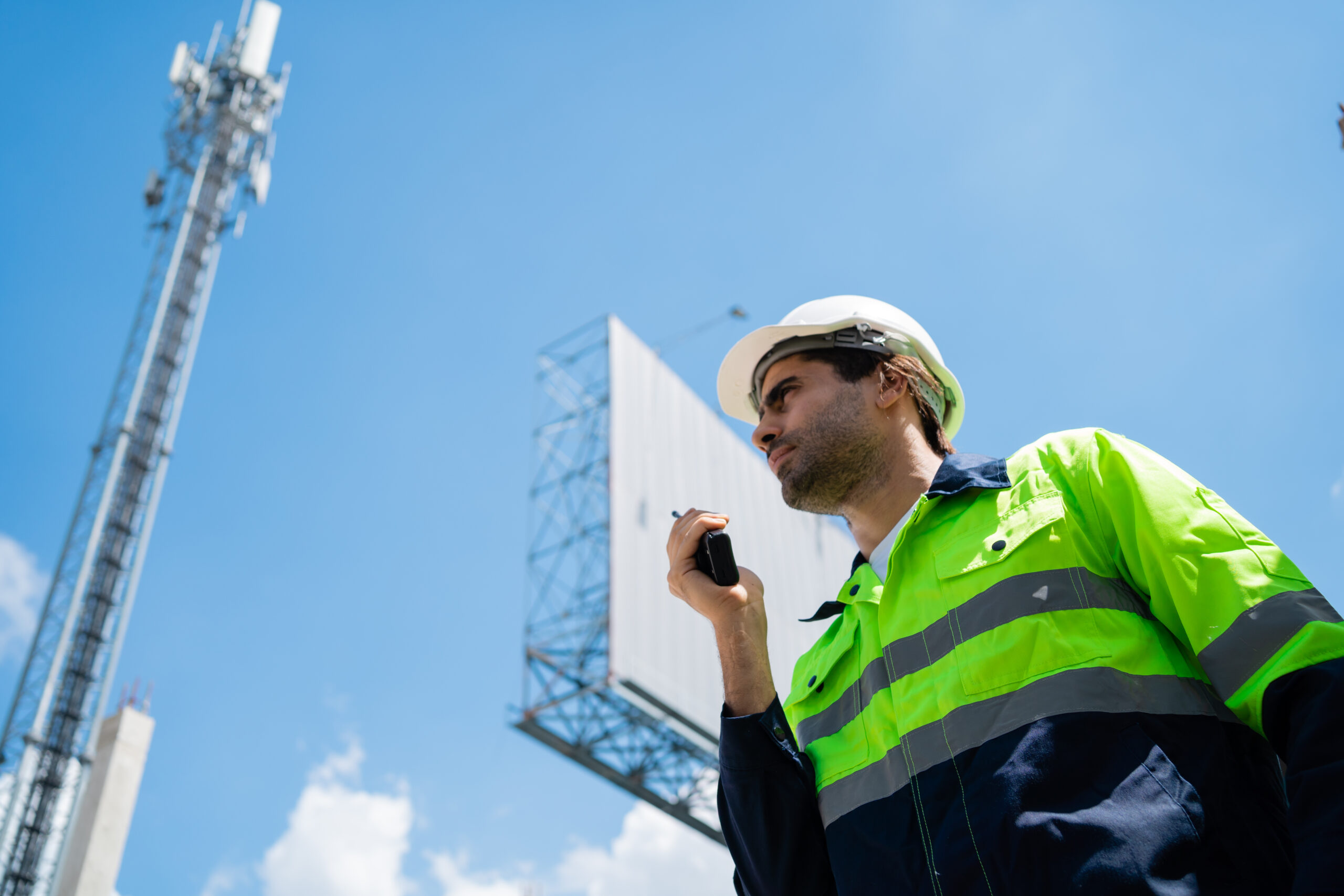 A man in a yellow and blue safety vest is talking on a cell phone. He is standing in front of a large billboard