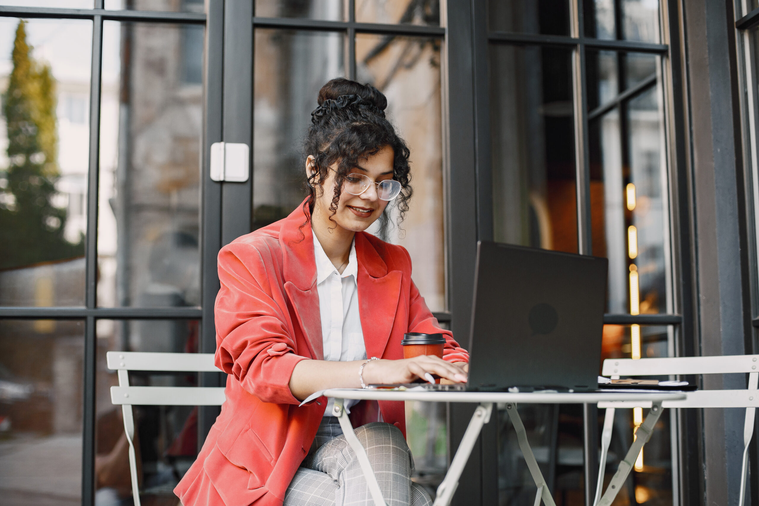Indian woman working on a laptop in a street cafe. Wearing stylish smart clothes - jacket, glasses