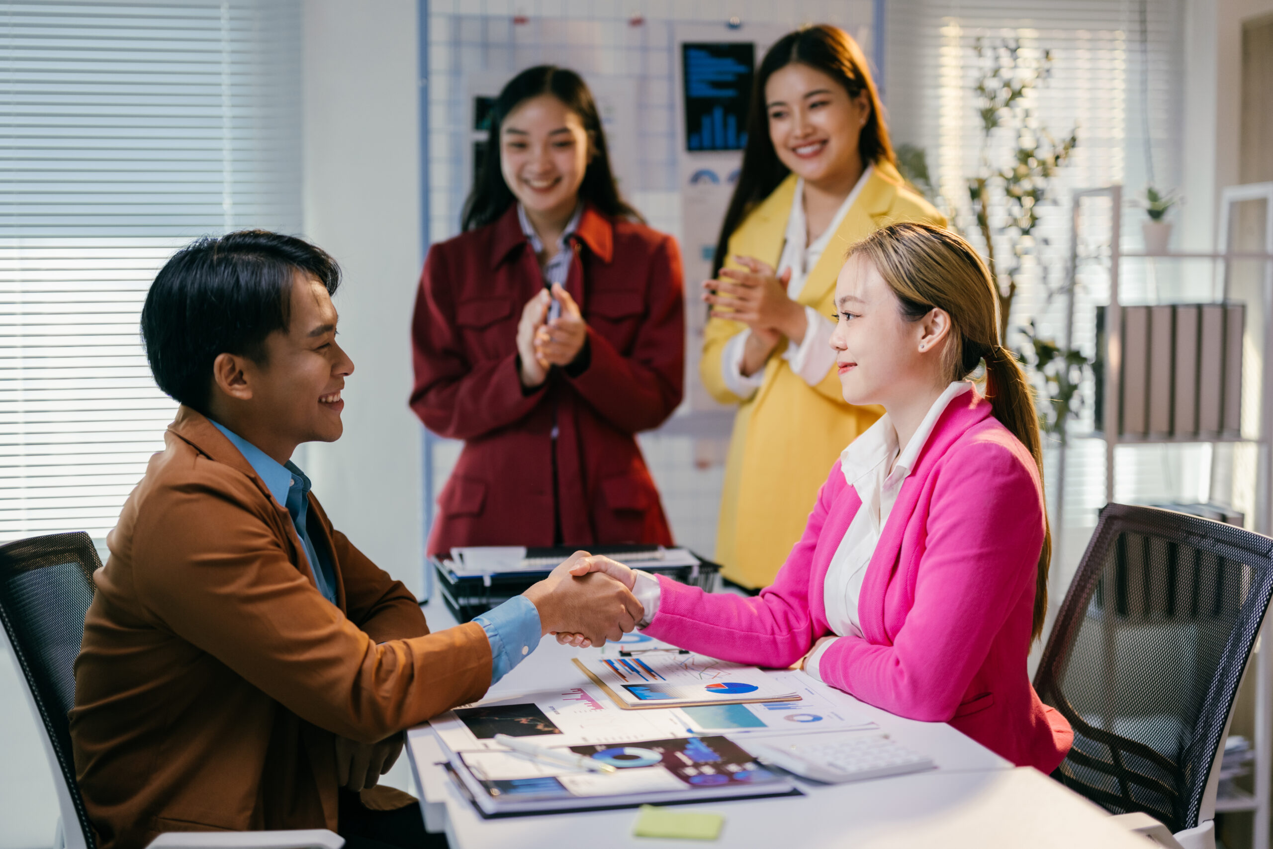Young businessman and businesswoman shaking hands after closing a deal in a modern office, their teammates are applauding them in the background