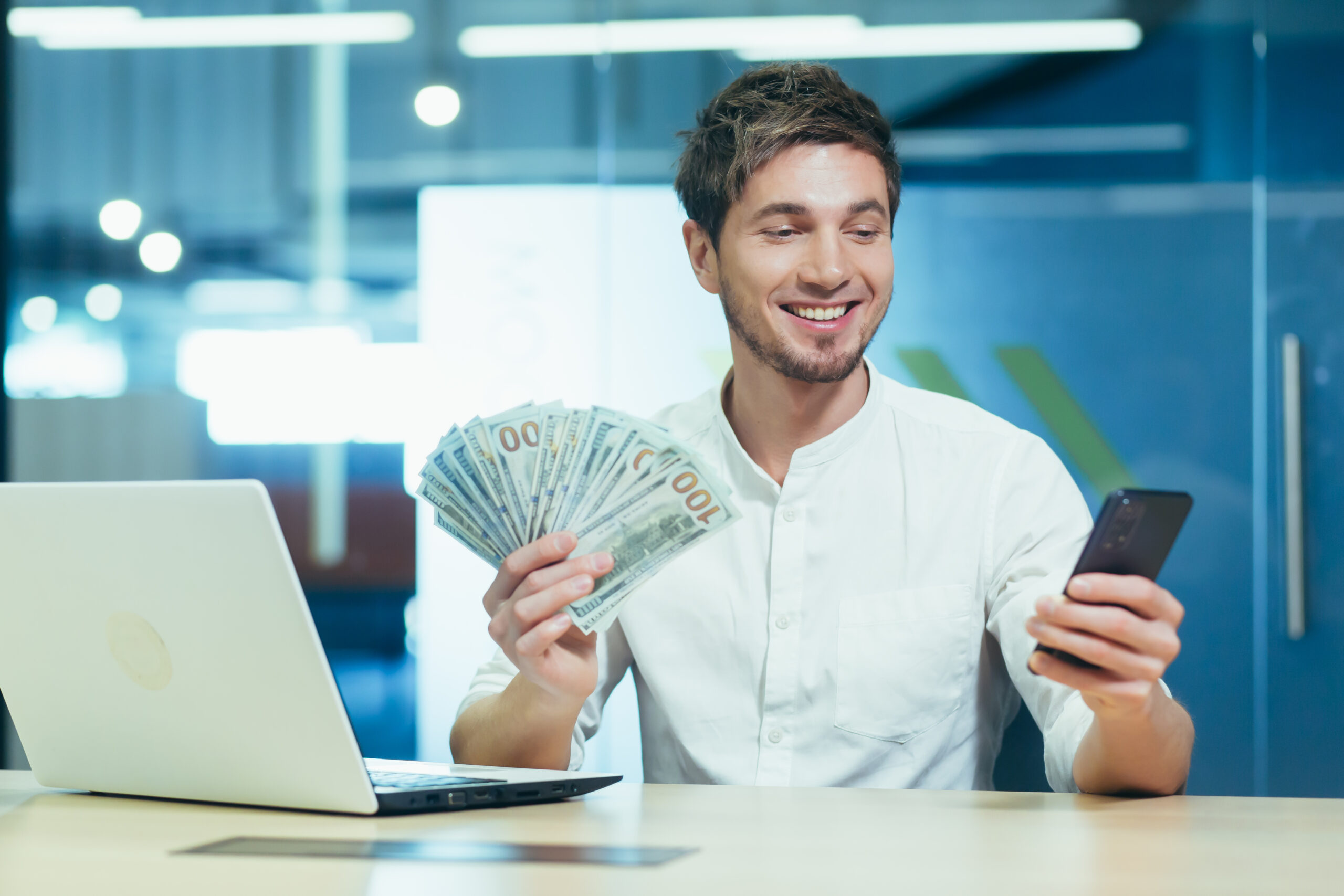 Young successful freelancer in the office holding in his hand winning banknote dollars, smiling and looking at the phone screen